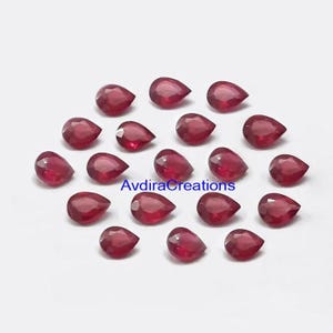 May include: A collection of faceted, teardrop-shaped, deep red gemstones. The gems are arranged on a white surface, with the text "AvdiraCreations" visible. These polished stones are suitable for jewelry making or crafting.