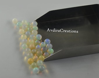 May include: A close-up of a pile of iridescent white opal beads in a metal scoop. The beads are round and have a slight shimmer to them. The text "AvdiraCreations" is visible in the background.