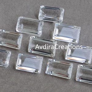 May include: A selection of clear, rectangular gemstones with a faceted cut. The gems are arranged on a grey surface, reflecting light and showcasing their transparent quality. The image features the text "AvdiraCreations".