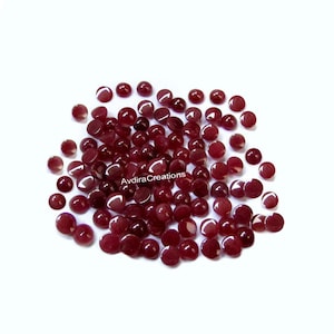 May include: A scattering of small, round, polished gemstones in a deep burgundy colour. The gemstones are spread across a white surface, reflecting light and creating highlights. The image is well-lit, showcasing the gems' smooth texture.