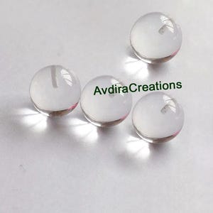 May include: Four clear glass marbles, each with a slight iridescent sheen. The marbles are arranged in a semi-circle on a white surface.