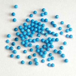 Op de afbeelding: Een verzameling kleine, ronde, turquoise blauwe cabochons. De cabochons zijn verspreid over een wit oppervlak. Avdira Creations staat in zwarte tekst op de afbeelding.