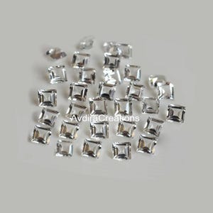 Op de afbeelding: Een verzameling heldere, vierkant geslepen strass-steentjes verspreid op een lichtgrijze ondergrond. De strass-steentjes zijn gefacetteerd en reflecteren licht. De tekst "AvdiraCreations" is zichtbaar.