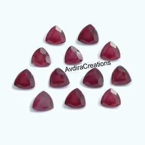 Pode incluir: Uma coleção de pedras preciosas triangulares, facetadas, vermelho escuro. As gemas estão dispostas sobre uma superfície branca. O texto "AvdiraCreations" é visível na imagem. Estas gemas são adequadas para fazer joias.