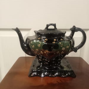 May include: A black ceramic teapot with a lid, spout, and handle. The teapot has a decorative floral pattern in green and gold. It sits on a matching black base, displayed on a brown wooden surface.