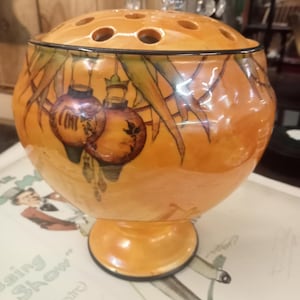 May include: A vintage orange ceramic flower frog. The round body sits on a pedestal base. The top has holes for flower arranging. The body is decorated with hand-painted orange lanterns and foliage.