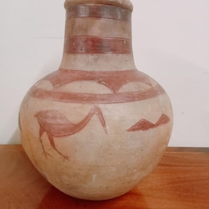 Könnte beinhalten: Ein bauchiges Keramikgefäß mit engem Hals und breitem Boden, das eine hellbraune Oberfläche mit rotbraunen bemalten Motiven aufweist, darunter eine stilisierte Vogelfigur und geometrische Muster, die den Körper und den Hals umschließen.