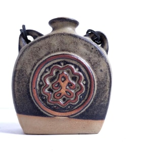 May include: A ceramic flask with a rounded body and a small neck. The flask is primarily tan with a dark brown band at the bottom and a decorative circular design in the center featuring orange and brown accents. It has two handles and a leather strap.