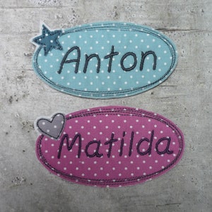 Könnte beinhalten: Zwei ovale Stoff-Namensschilder mit weißen Punkten. Das obere Schild ist hellblau mit einem blauen Stern und dem Namen "Anton" in Schwarz gestickt. Das untere Schild ist rosa mit einem grauen Herzen und dem Namen "Matilda" in Schwarz gestickt.
