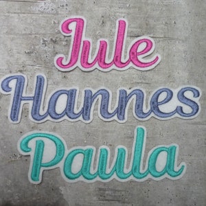 Könnte beinhalten: Drei gestickte Namen, Jule in Pink, Hannes in Blau und Paula in Türkis, auf grauem Hintergrund.