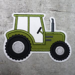 Könnte beinhalten: Gestickter Aufnäher eines grünen Traktors mit schwarzen Reifen und einer weißen Kabine. Der Traktor hat ein Lenkrad und einen Schornstein. Der Aufnäher hat einen weißen Rand und ist für Kleidung oder Bastelarbeiten geeignet.