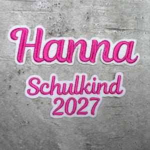 Könnte beinhalten: Ein weißer Stickaufnäher mit pinkfarbenen Nähten, der das Wort Hanna in Schreibschrift zeigt, gefolgt von dem Wort Schulkind und der Zahl 2027 in einer passenden pinken Schreibschrift, alles mit einem weißen Rand.