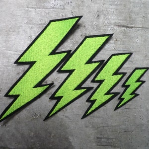 Könnte beinhalten: Vier neongrüne Blitz-Patches mit schwarzen Rändern sind diagonal auf einer grauen Oberfläche angeordnet. Die Patches variieren in der Größe, wobei das größte oben links und das kleinste unten rechts ist. Die Patches sind gestickt.