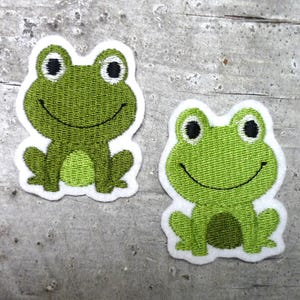 Könnte beinhalten: Zwei grüne gestickte Frosch-Patches mit weißen Umrandungen. Die Frösche haben schwarze Augen und einen lächelnden Mund.