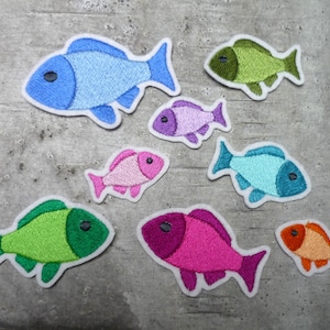 Könnte beinhalten: Acht bunte, gestickte Bügelbilder in Fischform. Die Patches sind in Blautönen, Grün, Rosa und Orange.