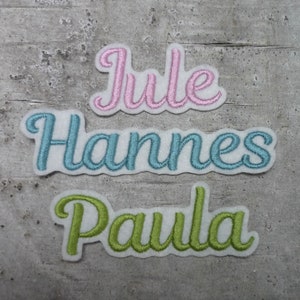 Könnte beinhalten: Drei gestickte Stoff-Patches mit Namen in Schreibschrift. Der obere Patch ist rosa und sagt "Jule". Der mittlere Patch ist blau und sagt "Hannes". Der untere Patch ist grün und sagt "Paula".
