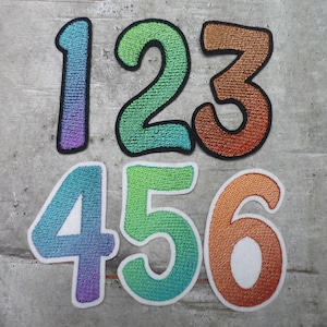 Könnte beinhalten: Sechs bunte gestickte Bügelbilder in Form der Zahlen 1 bis 6. Die Zahlen sind schwarz umrandet und haben einen Farbverlaufseffekt. Die Zahlen sind in zwei Reihen zu je drei angeordnet.