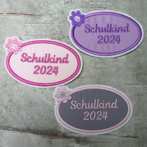 Könnte beinhalten: Drei ovale Stoffpatches mit rosa, lila und grauem Hintergrund. Jeder Patch hat einen weißen Rand und eine rosa Blume in der oberen linken Ecke. Der Text "Schulkind 2024" ist in weiß auf jedem Patch gestickt.