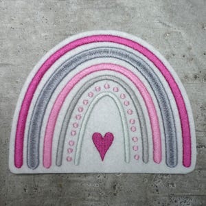 Könnte beinhalten: Ein weißer Filz-Regenbogen-Patch mit rosa und grauen gestickten Streifen und einem rosa Herz in der Mitte.