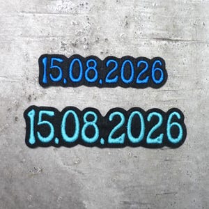 Könnte beinhalten: Zwei schwarze Stoffaufnäher mit dem gestickten Datum 15.08.2026. Der obere Aufnäher zeigt das Datum in königsblauem Garn, während der untere Aufnäher dasselbe Datum in einem helleren Cyanblau mit einer dickeren Schriftart darstellt.