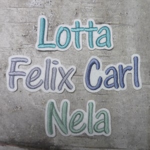 Könnte beinhalten: Vier gestickte Namensaufnäher: "Lotta" in Türkis, "Felix" in Lavendel, "Carl" in Blau und "Nela" in Mintgrün. Jeder Name ist auf weißem Hintergrund mit einer verspielten, abgerundeten Schrift gestickt. Die Aufnäher sind auf einer strukturierten, grauen Oberfläche angeordnet.
