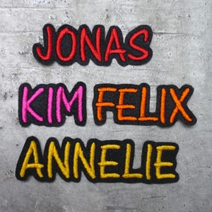 Könnte beinhalten: Vier gestickte Patches mit Namen darauf. Die Namen sind Jonas, Kim, Felix und Annelie. Die Patches befinden sich auf einem grauen Hintergrund.