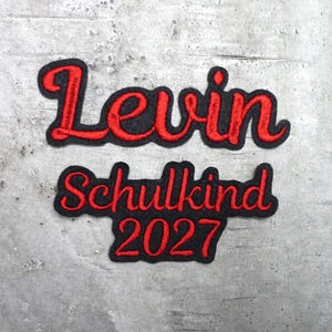 Könnte beinhalten: Ein schwarzer Stickaufnäher mit roter Bestickung, der den Namen Levin, das Wort Schulkind und das Jahr 2027 zeigt. Der Text ist in einer Schreibschrift gehalten und von einem dicken schwarzen Rand um die roten Buchstaben umgeben.