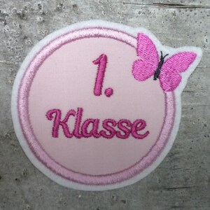 Könnte beinhalten: Rosa-weißer gestickter Aufnäher mit einem rosa Schmetterling und dem Text "1. Klasse".