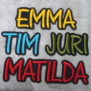 Könnte beinhalten: Vier gestickte Aufnäher mit Namen in verschiedenen Farben. Die Namen sind Emma in Gelb, Tim in Blau, Juri in Grün und Matilda in Rot.