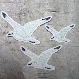 Könnte beinhalten: Drei weiße gestickte Möwen-Patches mit schwarzen Umrissen. Die Patches befinden sich auf einem grauen, strukturierten Hintergrund.