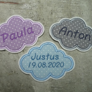 Könnte beinhalten: Drei personalisierte, wolkenförmige Stoff-Patches mit weißen Punkten. Die Patches sind in Lila, Grau und Blau. Der lila Patch sagt "Paula", der graue Patch sagt "Anton" und der blaue Patch sagt "Justus 19.08.2020".