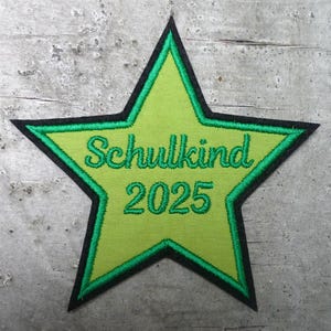 Könnte beinhalten: Ein grüner sternförmiger Patch mit schwarzem Rand. Der Patch ist mit dem Text "Schulkind 2025" in grünem Garn bestickt.