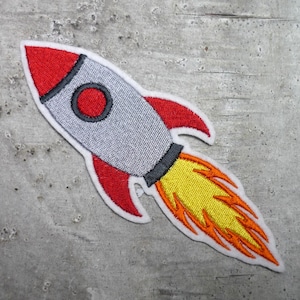 Könnte beinhalten: Ein gestickter Raketen-Patch in Rot, Grau und Weiß mit einer gelben und orangenen Flamme. Die Rakete fliegt nach oben.