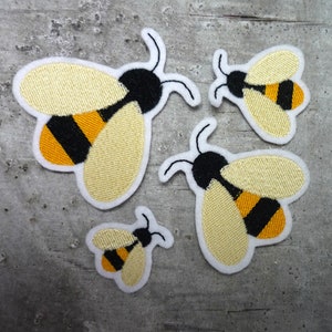Könnte beinhalten: Vier gestickte Bienen-Patches in verschiedenen Größen. Die Bienen sind gelb mit schwarzen Streifen und haben schwarze Augen. Die Patches sind weiß mit einem schwarzen Umriss.