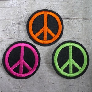 Könnte beinhalten: Drei Peace-Zeichen-Patches in verschiedenen Farben. Die Patches sind schwarz mit einem weißen Peace-Zeichen in der Mitte. Die Peace-Zeichen sind in Orange, Pink und Grün umrandet.