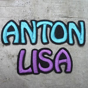 Könnte beinhalten: Zwei gestickte Patches mit den Namen "ANTON" in Blau und "LISA" in Lila auf einem grauen, strukturierten Hintergrund.
