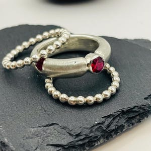 Op de afbeelding: Drie zilveren ringen met een rode edelsteen op de middelste ring. De ringen zijn gestapeld op een donkergrijs oppervlak.