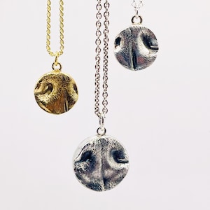 May include: Three dog nose pendant necklaces. One gold-coloured pendant on a gold chain, and two silver-coloured pendants on silver chains. Each pendant is a circular shape with a detailed dog nose design.