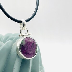May include: A round pendant with a faceted, deep magenta gemstone, set in a silver bezel. The pendant hangs from a black cord. The gemstone has green inclusions. The pendant is displayed against a light blue background.