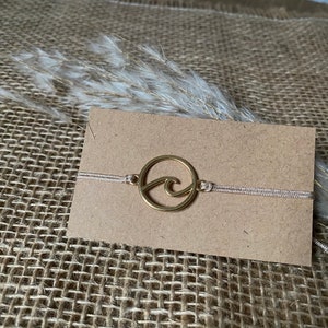Könnte beinhalten: Ein goldfarbenes Armband mit einem Wellen-Anhänger auf einer beigefarbenen Schnur. Das Armband wird auf einer braunen Karte präsentiert.