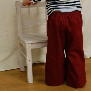 Puede incluir: Un ni&ntilde;o con un pantal&oacute;n rojo de piernas anchas y una camisa a rayas de pie frente a una silla de madera blanca.