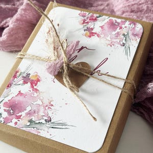 Op de afbeelding: Een bruine kartonnen doos met een witte kaart met aquarel bloemenkunst in roze en rode tinten. De doos is vastgebonden met touw en versierd met een klein bruin hart en gedroogde bloemen. De achtergrond is een zachte roze stof.