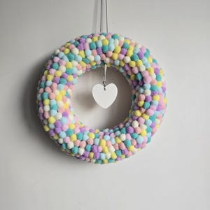 Könnte beinhalten: Ein pastellfarbener Kranz aus kleinen, runden, bunten Kugeln in Rosa, Gelb, Blau und Lila. In der Mitte des Kranzes hängt ein weißes herzförmiges Ornament an einem dünnen Faden.