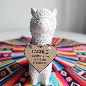 Könnte beinhalten: Eine weiße Alpaka-Figur aus Keramik mit strukturiertem Fell, die ein herzförmiges Holzschild trägt. Auf dem Schild steht der deutsche Text "LÄCHLE! Du kannst sie nicht alle anspucken!". Das Alpaka steht auf einem bunten, gemusterten Textil.