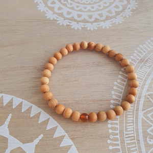 Könnte beinhalten: Ein Armband aus Holzperlen mit einer einzigen orangefarbenen Perle. Das Armband liegt auf einer hellbraunen Holzoberfläche mit weißen Mandala-Mustern.