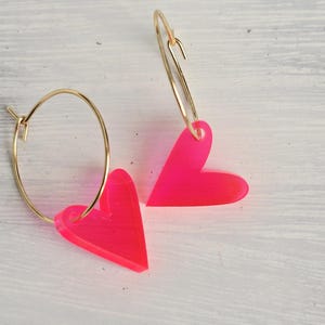 Könnte beinhalten: Goldene Creolen-Ohrringe mit leuchtend pinkfarbenen herzförmigen Anhängern. Die Ohrringe sind auf einer weißen Holzoberfläche platziert, was einen Kontrast zur leuchtenden Farbe der Herzen bildet.