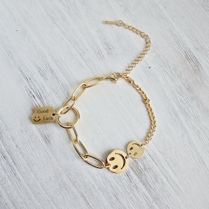 Könnte beinhalten: Goldfarbenes Armband mit einer Mischung aus Kettenstilen und Smiley-Anhängern. Ein Anhänger trägt die Aufschrift "Good Luck" mit einem lächelnden Gesicht. Das Armband hat einen Karabinerverschluss und wird auf einer weißen Holzoberfläche präsentiert.