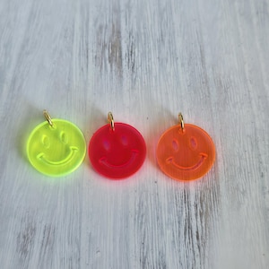 Könnte beinhalten: Drei kreisförmige Acryl-Anhänger mit einem Smiley-Gesicht-Design. Die Anhänger sind in Neongelb, Neonpink und Neonorange gehalten und jeweils mit einem kleinen goldfarbenen Metallring an der Oberseite zum Aufhängen oder Verbinden versehen.