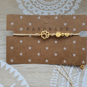 Könnte beinhalten: Ein handgemachtes beigefarbenes Armband aus Schnur mit goldenen Anhängern, darunter ein Pfotenabdruck, ein Buchstabe A, eine Perle und ein Herz. Das Armband wird auf einer braunen Karte mit weißen Sternen und dem Wort "HANDMADE" präsentiert.