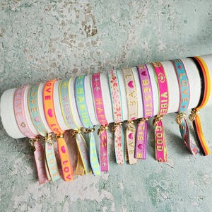 Könnte beinhalten: Eine Sammlung von elf farbenfrohen Stoffarmbändern mit verschiedenen Mustern und Texten wie LOVE, VIBES, HAKUNA, HAPPINESS und BESTIE. Jedes Band hat einen kleinen goldfarbenen Metallanhänger und endet in lose herabhängenden Stoffenden.